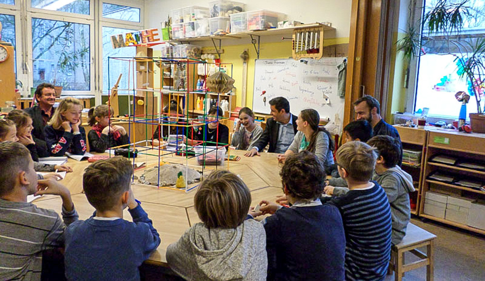 Foto: Kinder und Erwachsene sitzen an einen große runden Tisch mit einer Konstruktion darauf