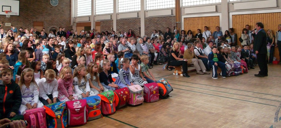 Foto: Kinder mit Eltern sitzend auf Bänken in der Turnhalle zur Einschulung. Mann mit Anzug hält Ansprache.