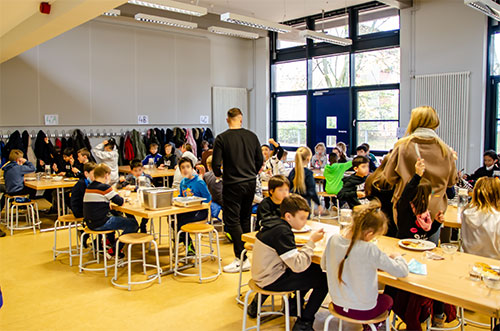 Foto: Kinder sitzen an Tischen und essen, dazwischen stehend Lehrer und Erzieher