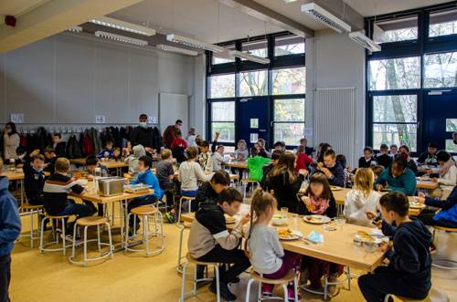 Foto: Jungen und Mädchen sitzen an gruppierten Tischen und essen in einem hohen Raum mit Fensterfront im Hintergrund